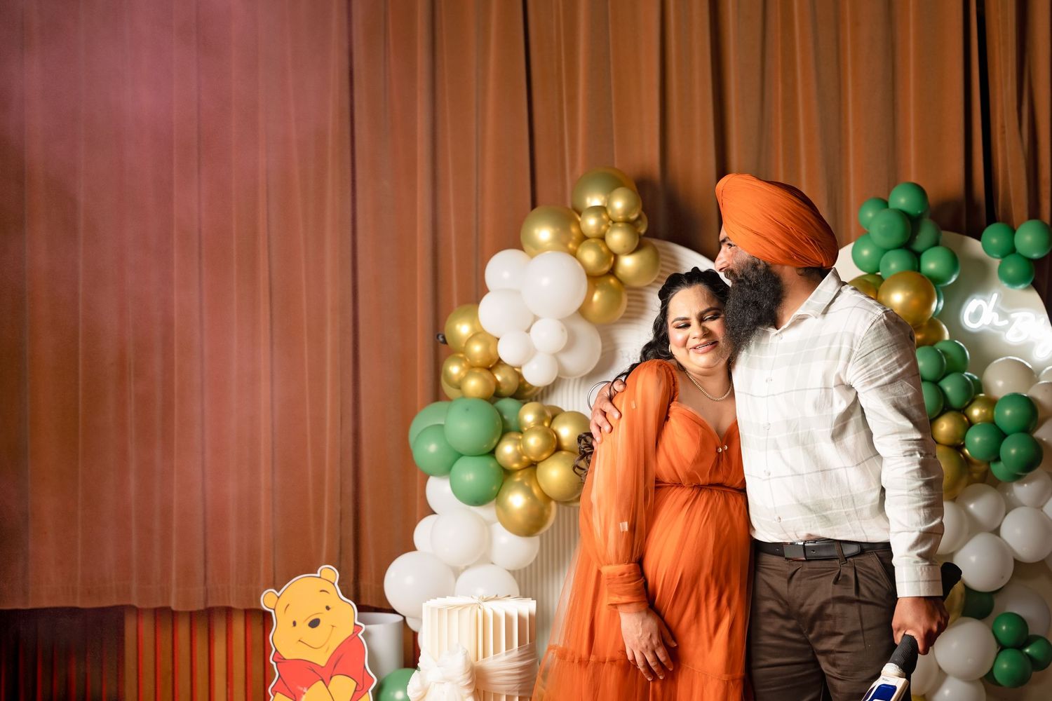 Couple embracing at Winnie the Pooh themed baby shower with green and gold balloon arch backdrop.