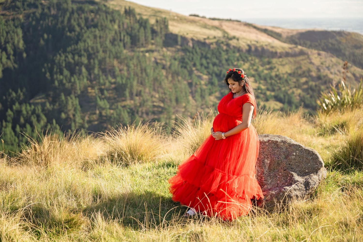 Pregnant woman in red gown poses on rocky hillside with lush green mountains in background.
