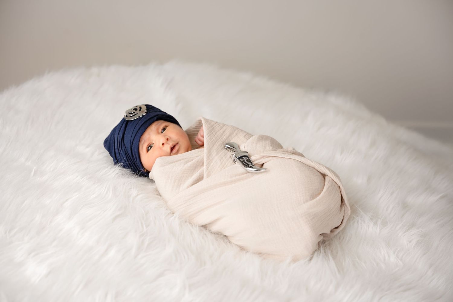 Newborn baby swaddled in cream wrap wearing navy blue hat, peacefully sleeping on white fluffy blanket.