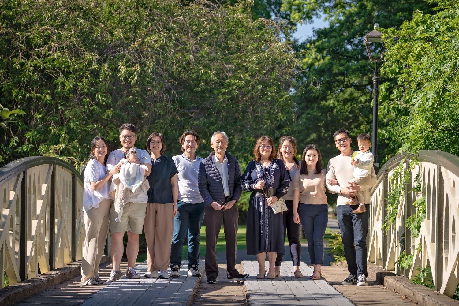 A group of people stand together smiling on a wooden footbridge surrounded by green trees on a sunny day.