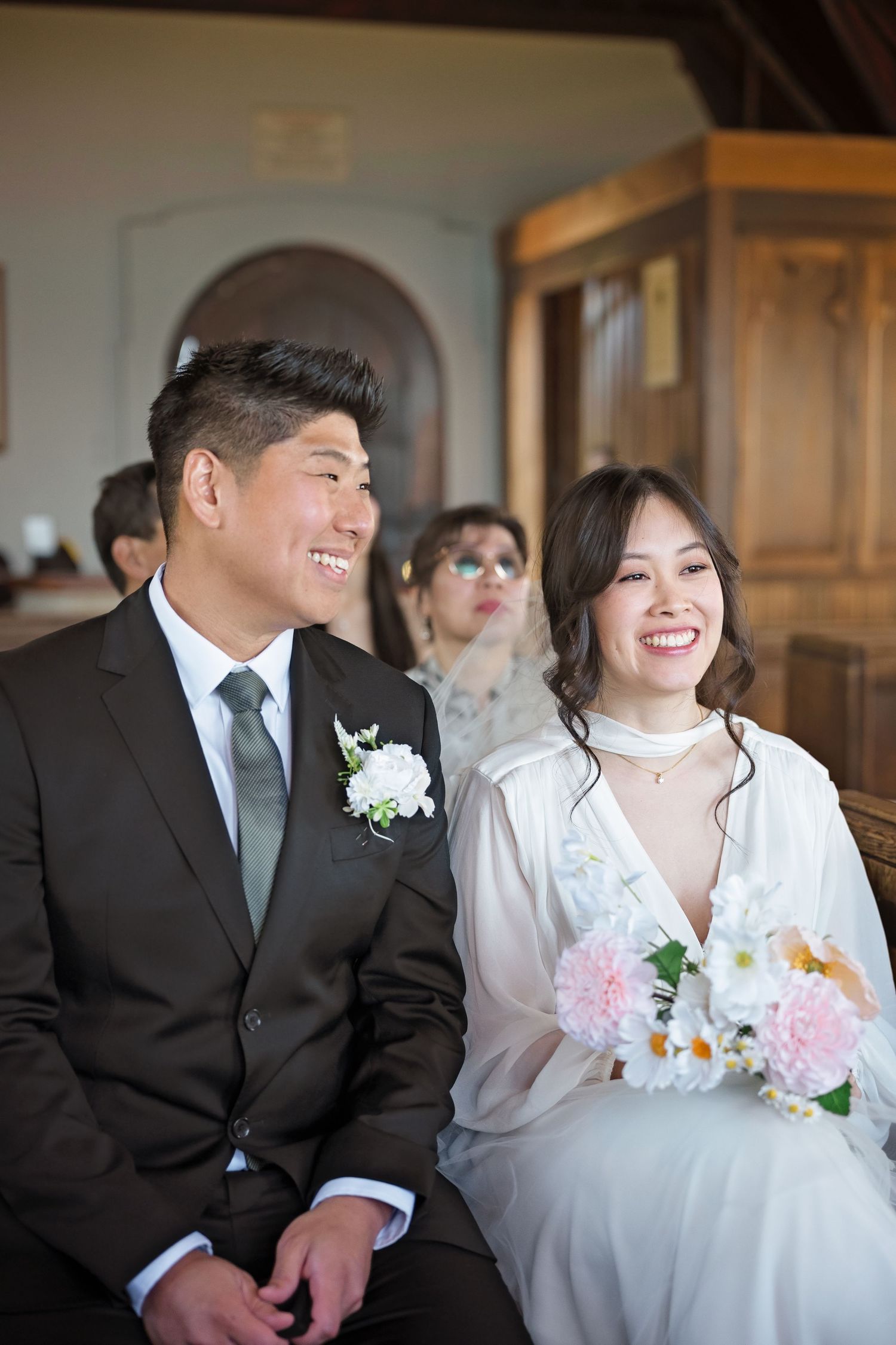 Smiling Asian bride holding pink flowers and groom in dark suit laugh joyfully during their wedding ceremony.