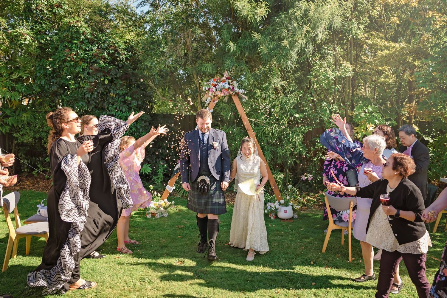 Wedding guests toss confetti as bride and groom walk through floral arch in sunny garden.