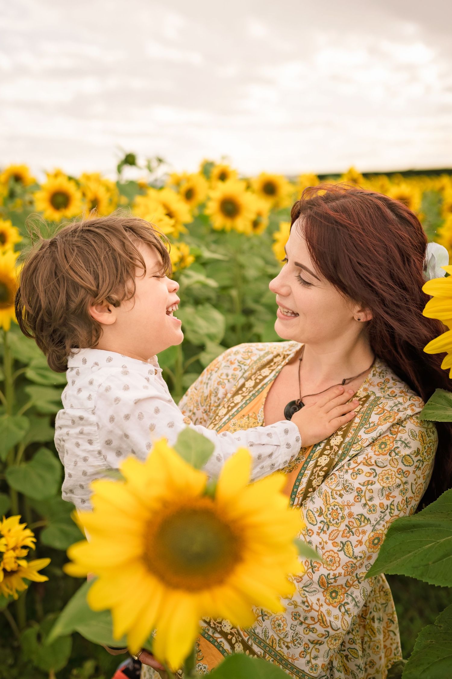 A joyful mother holds her laughing child in a vibrant sunflower field, both smiling warmly at each other.