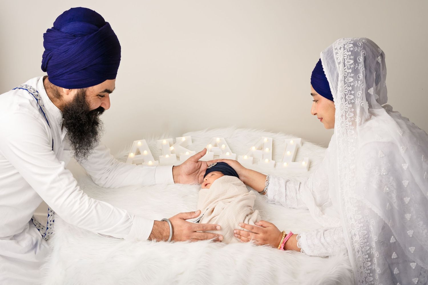 Sikh couple in white attire and blue turbans tenderly caring for their newborn baby during a family portrait session.