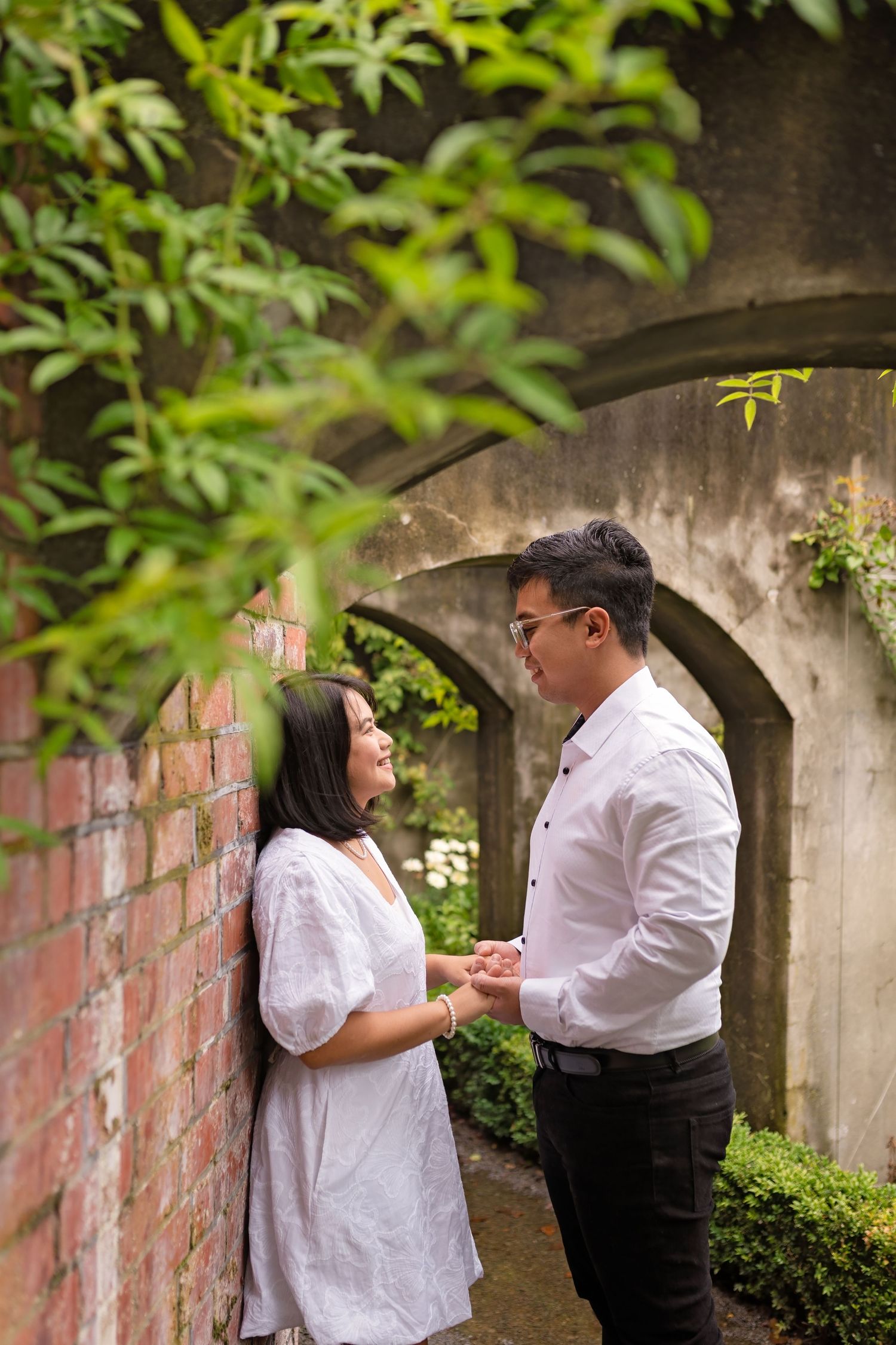 Couple sharing a romantic moment by a brick wall with arched stone architecture and lush green foliage.