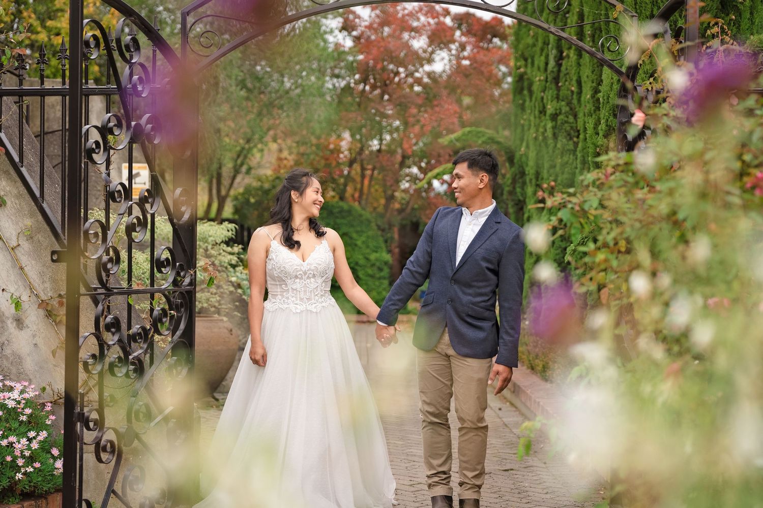 Bride and groom holding hands walking through an ornate iron gate surrounded by colorful garden blooms.