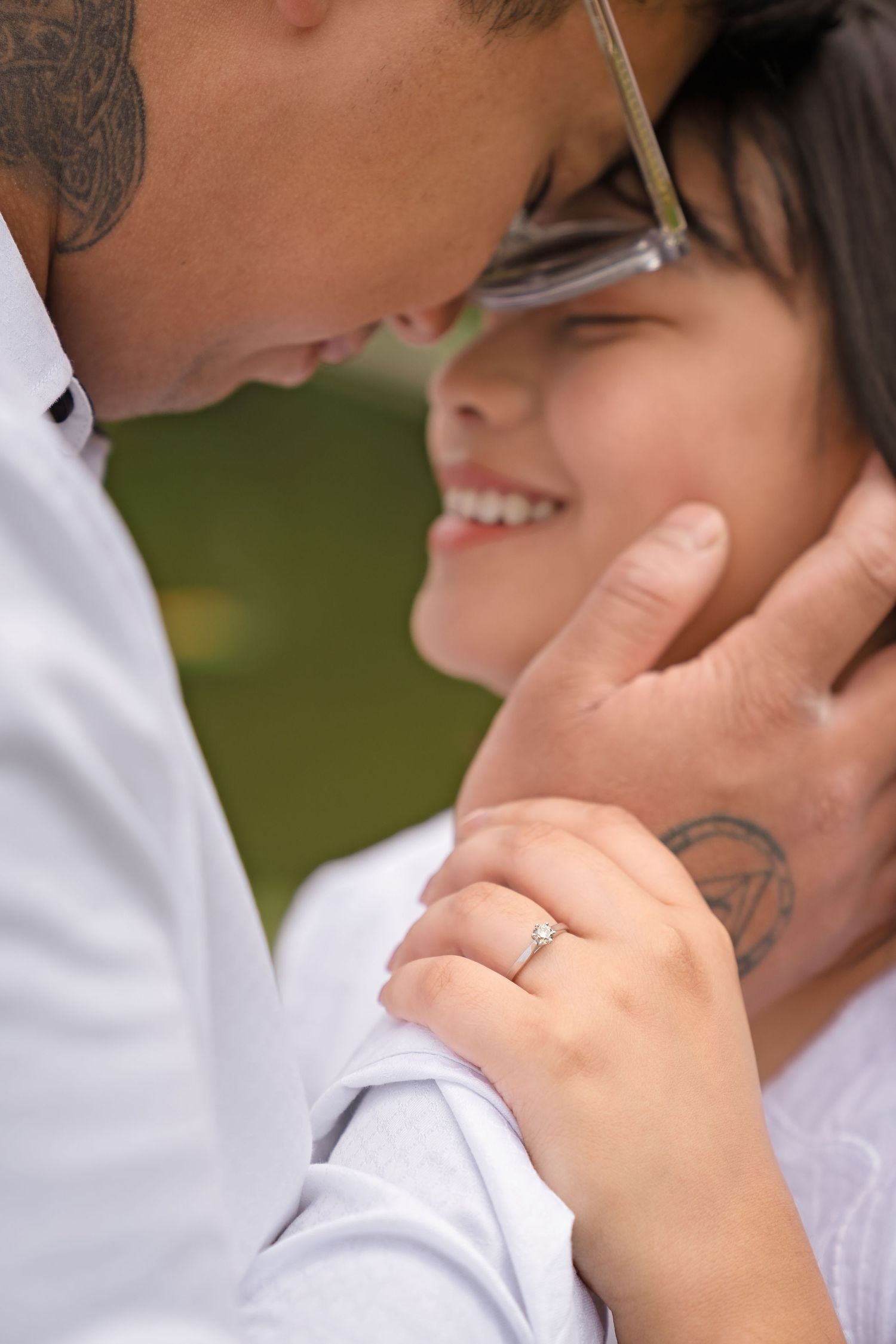Couple sharing a romantic moment, woman smiling with engagement ring visible, man with tattoos gently holding her face.