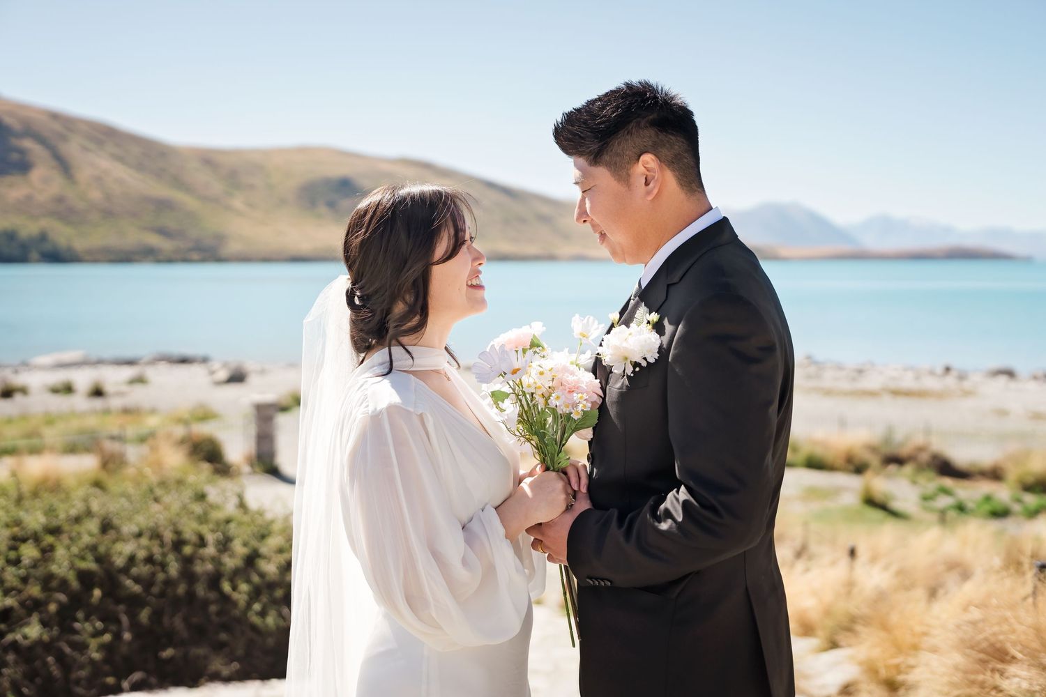 Bride and groom share an intimate moment holding white flowers with turquoise lake and mountains in background.