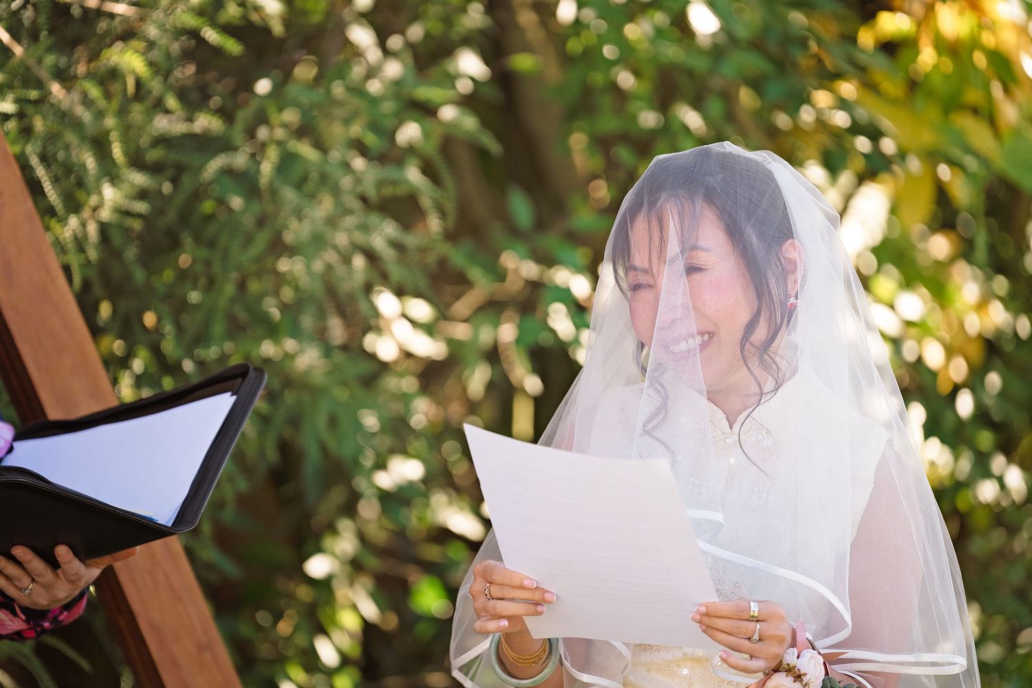 Smiling bride in white veil reads wedding vows from paper during outdoor garden ceremony.