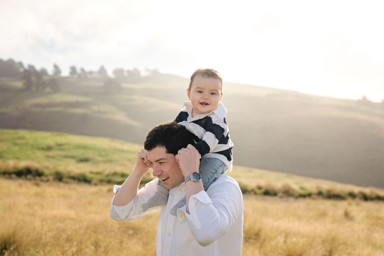 Father carrying smiling baby on shoulders in sunny green meadow, enjoying outdoor family moment together.