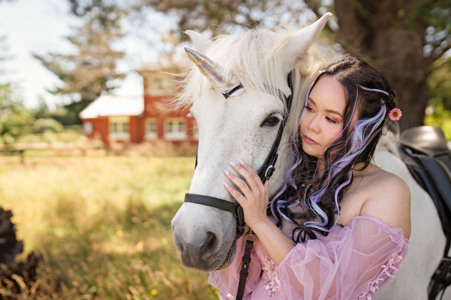 Young woman in pink dress with purple hair extensions tenderly embracing a white horse outdoors near a red barn.