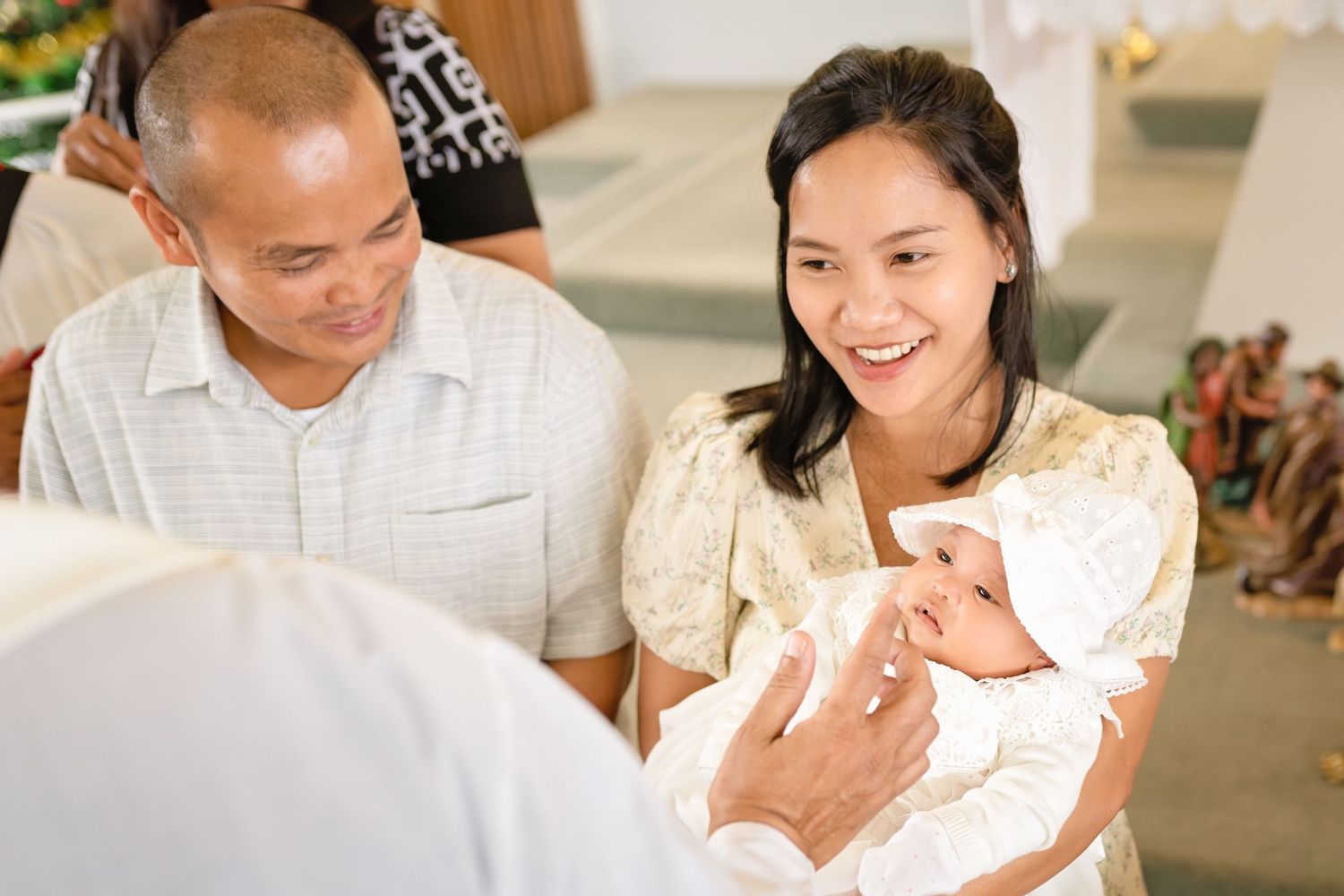 Asian couple smiling at a baby baptism ceremony in a church, mother holding newborn in white christening gown.