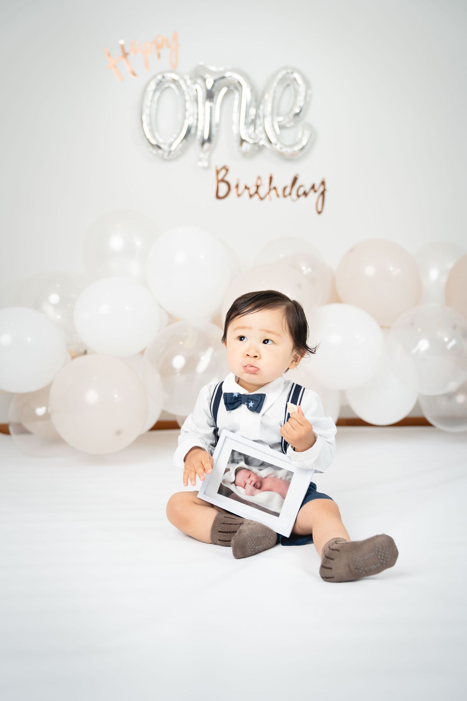 A toddler in suspenders and bow tie holds a photo frame during a first birthday photoshoot.