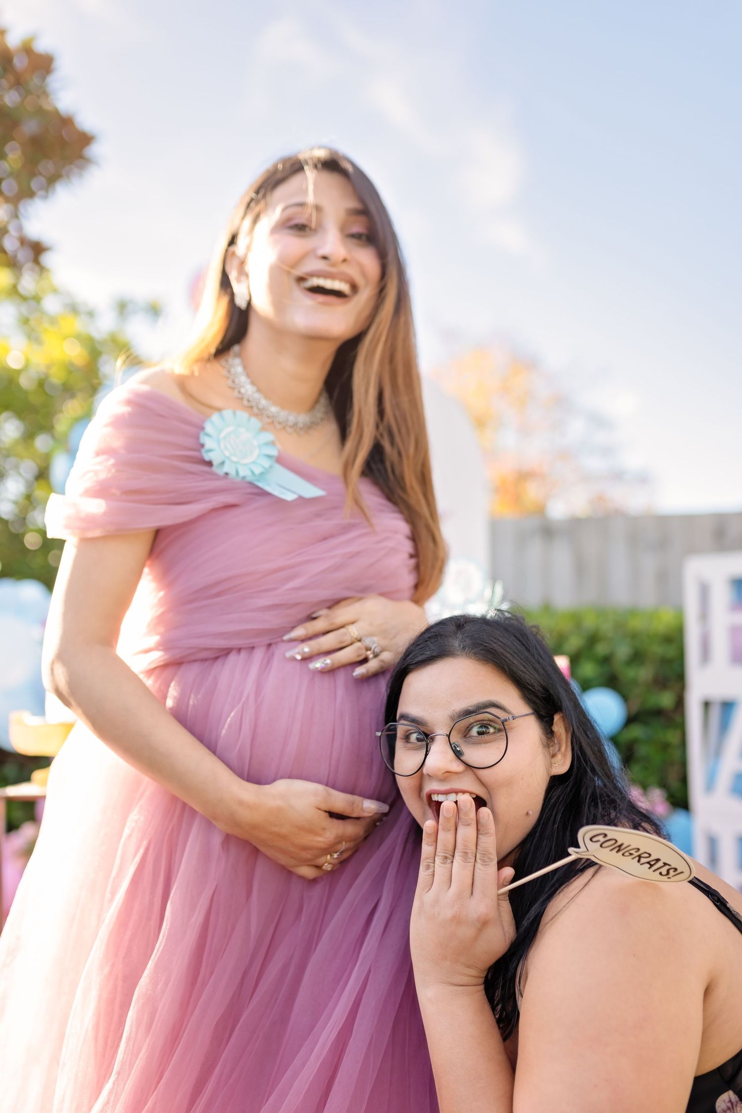 Two women in an outdoor photoshoot, one wearing a pink dress while the other playfully gestures during a celebratory moment.