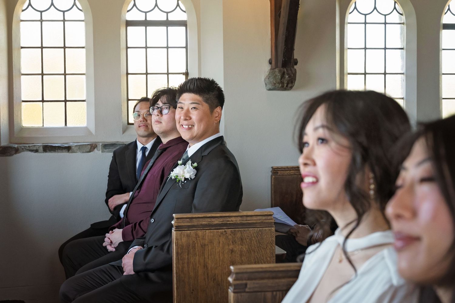 Wedding guests smile and laugh during a church ceremony seated in wooden pews with arched windows.