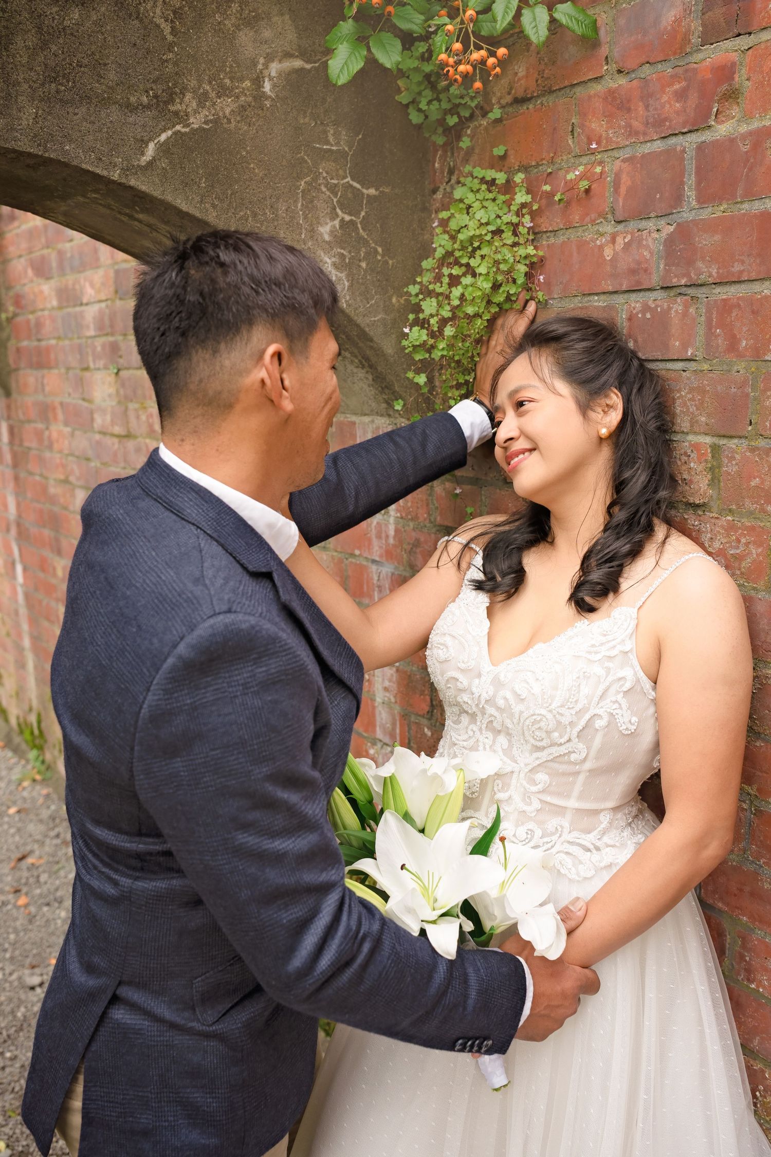 A groom in a gray suit tenderly touches his smiling bride's face as she holds white lilies against a brick wall.