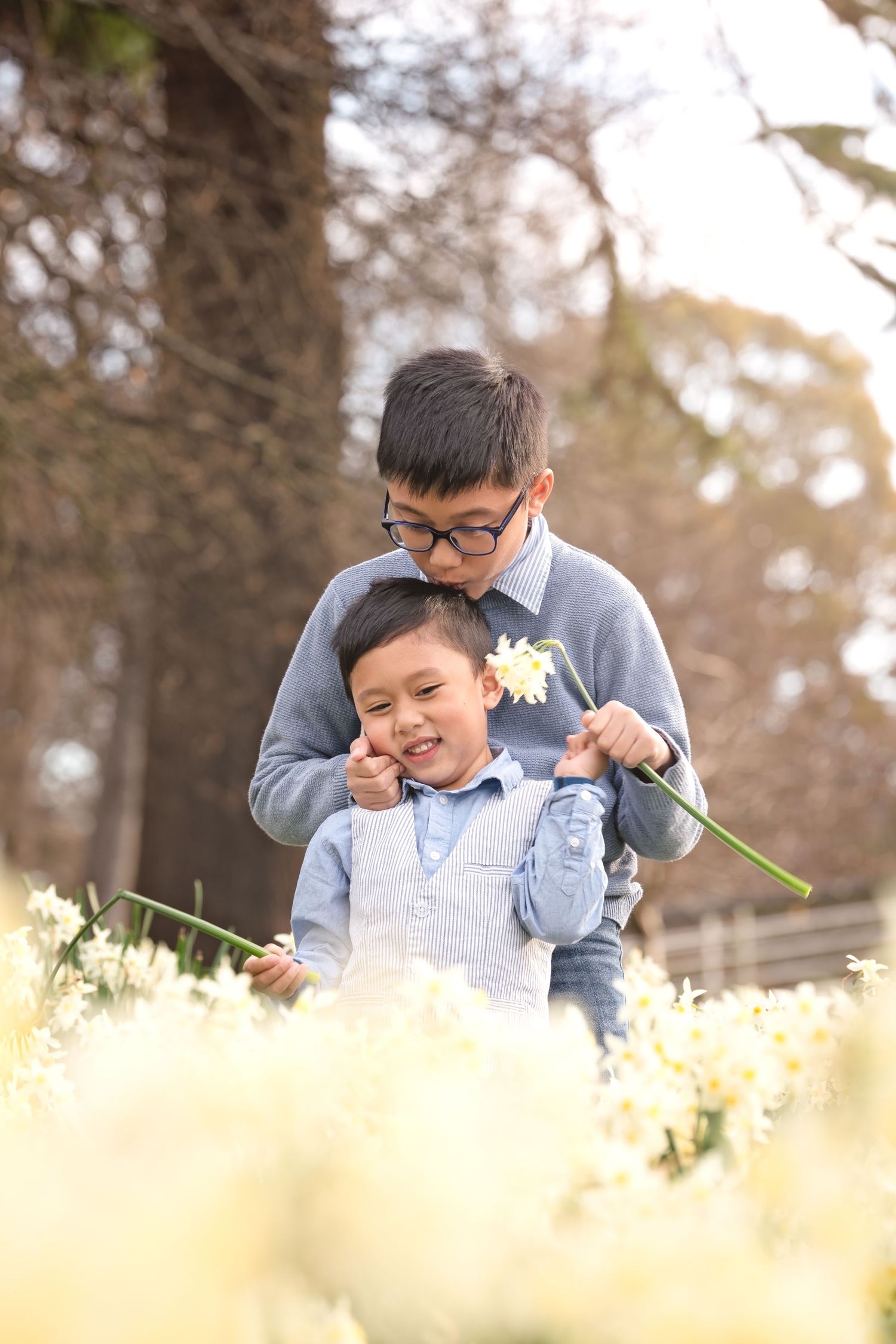 Two young boys playing among white daffodils in a park, older brother wearing glasses hugging younger sibling.
