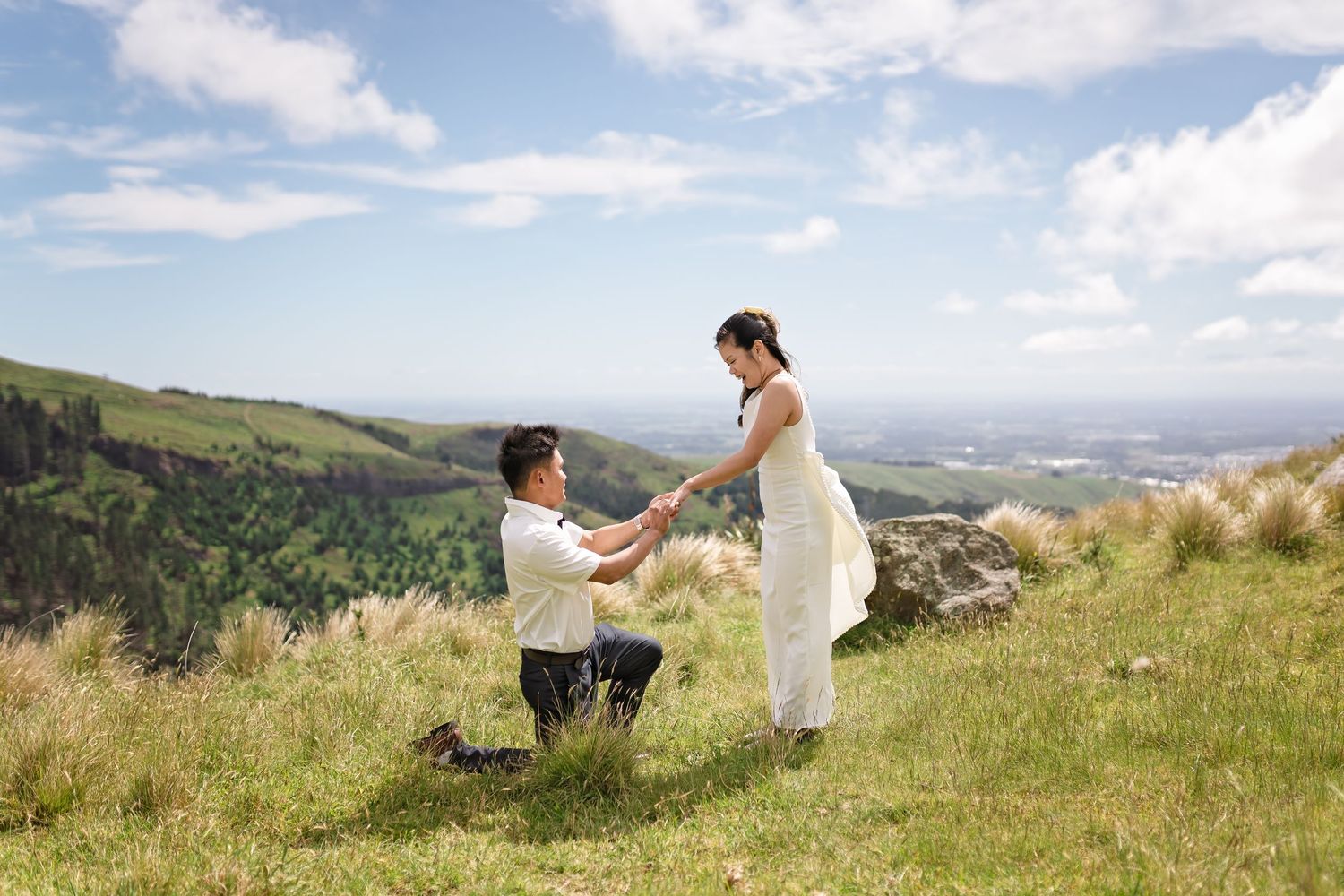 Man kneeling on grassy hilltop proposing to woman in white dress with scenic mountain landscape backdrop.