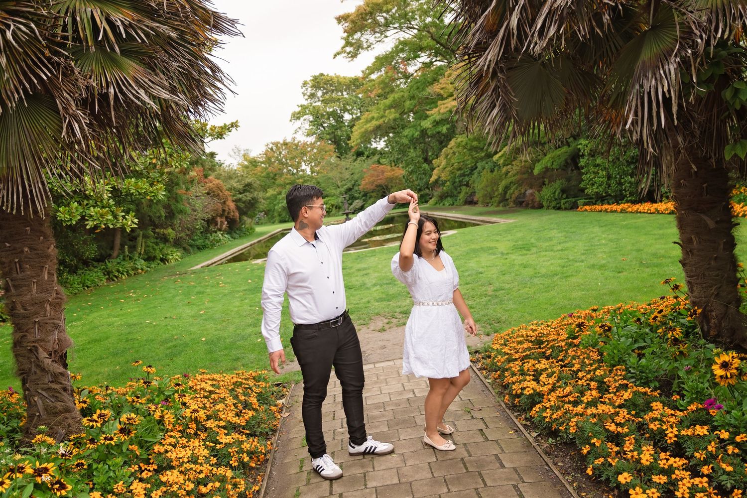 Couple dancing on a garden path surrounded by orange flowers and palm trees in a lush green park.