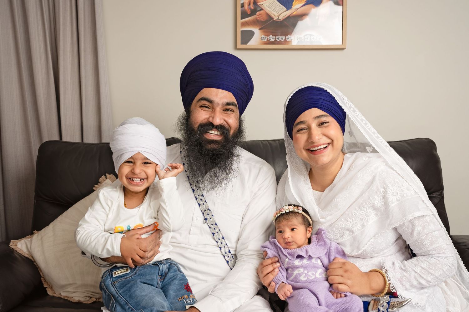 Sikh family smiling together on sofa, parents in traditional white attire and blue turbans, with toddler and newborn baby.