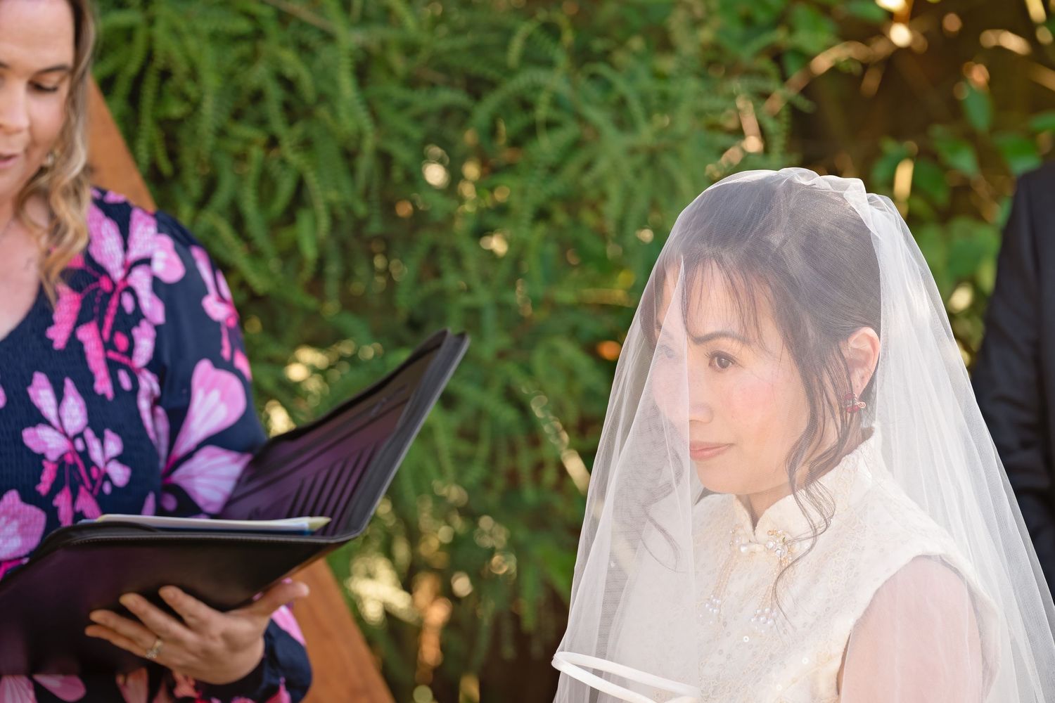 Bride wearing a white veil listens during outdoor wedding ceremony while officiant reads from a black folder.