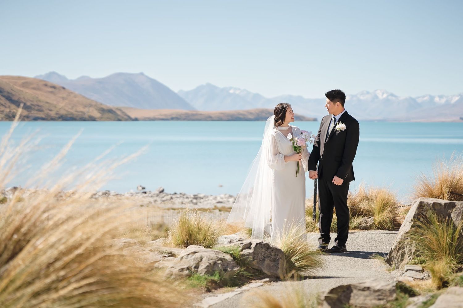 Bride and groom stand together on a lakeside path with turquoise water and snow-capped mountains in New Zealand.