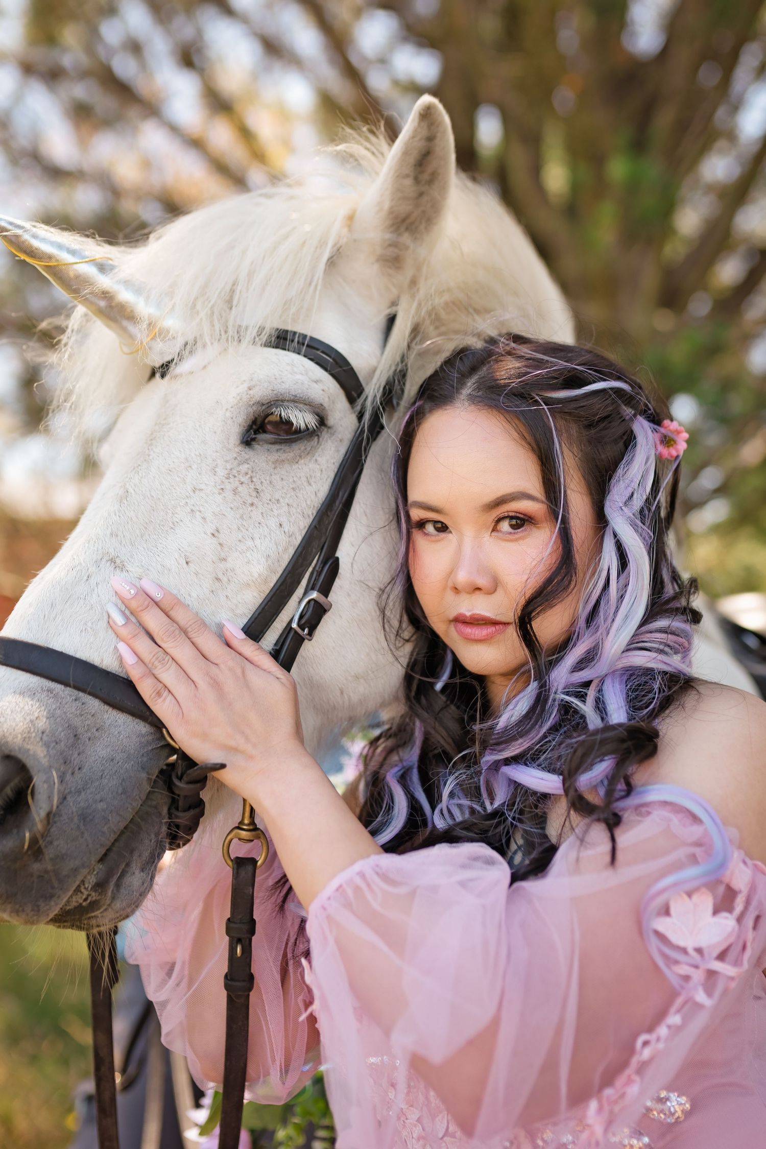 A woman in a pink dress with purple-highlighted hair gently leans against a white unicorn horse outdoors.