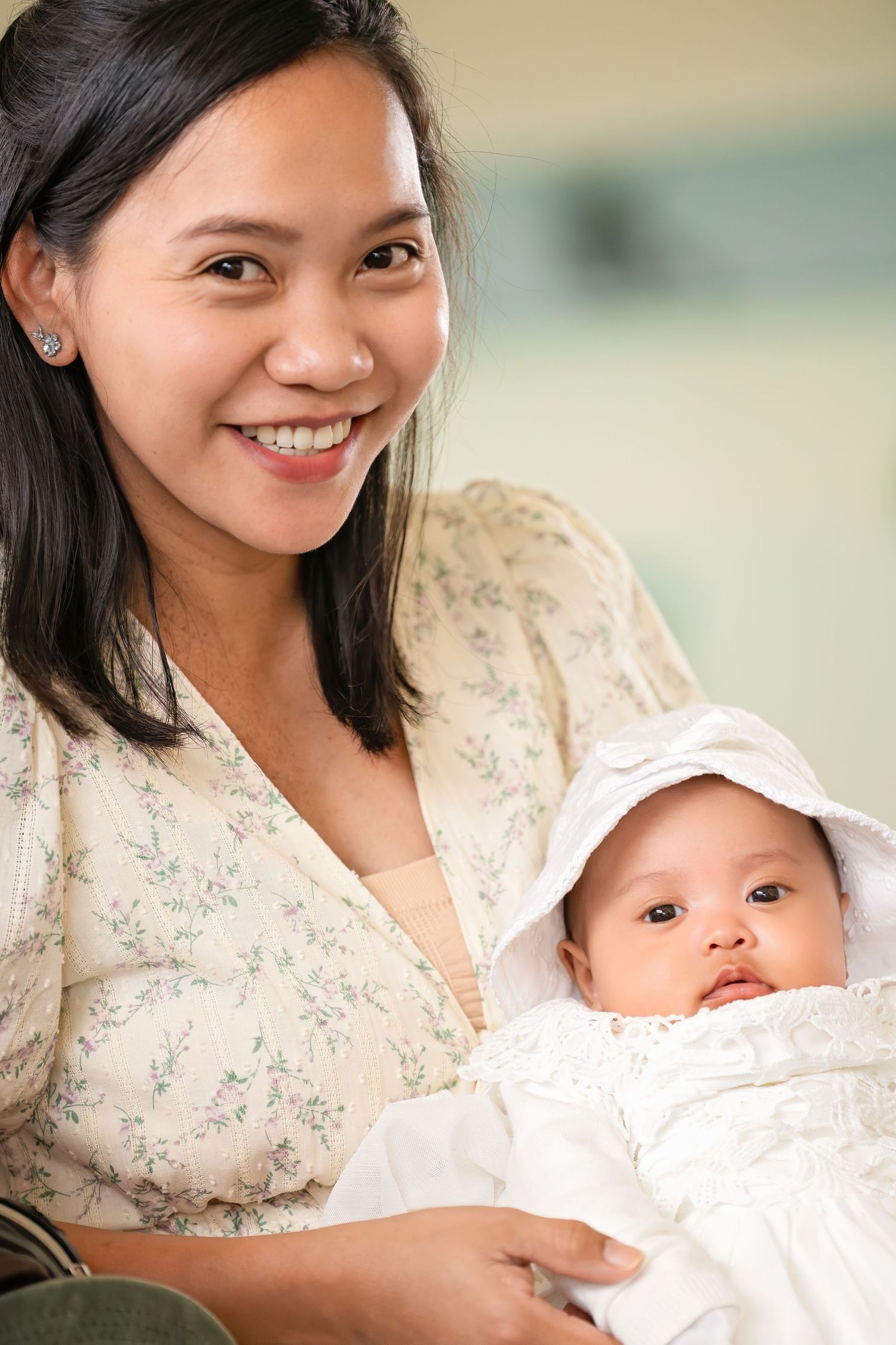 Smiling Asian mother holding her infant dressed in white christening gown and bonnet.