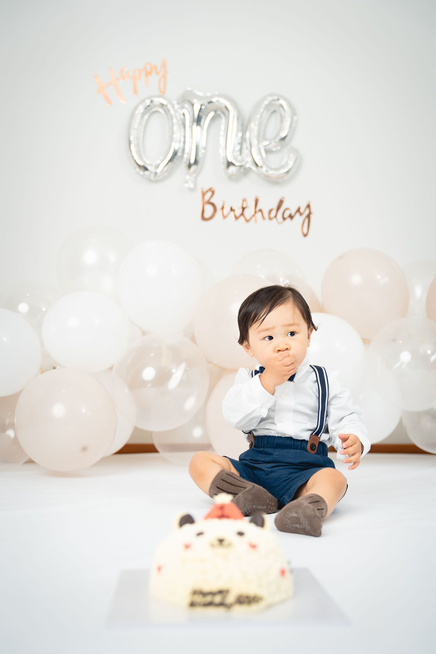 A small child sits on the floor with a birthday cake and silver 'ONE' balloon decorations against white balloons.