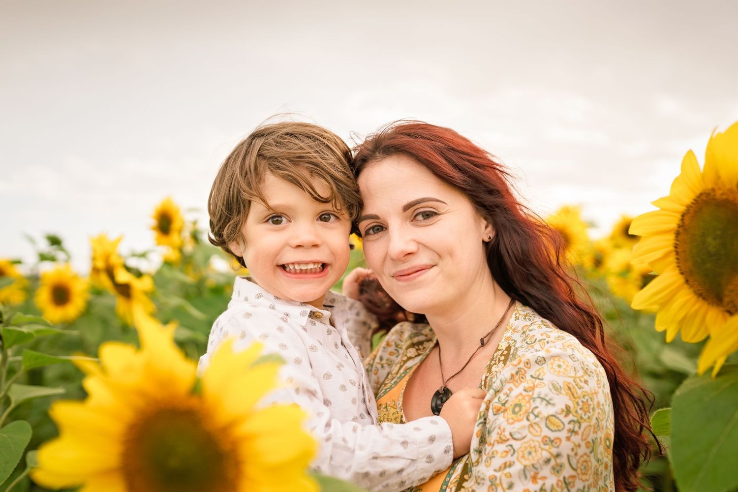 Woman with red hair holds smiling toddler in sunny sunflower field, both wearing floral outfits.
