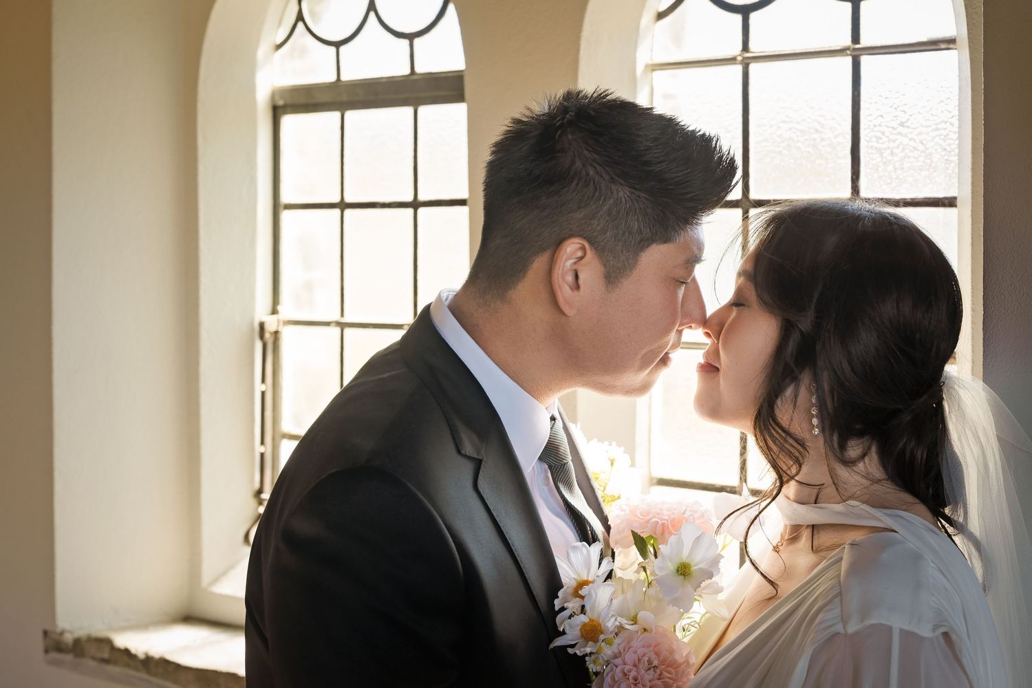 Asian bride and groom sharing a romantic kiss by sunlit windows holding a bouquet.