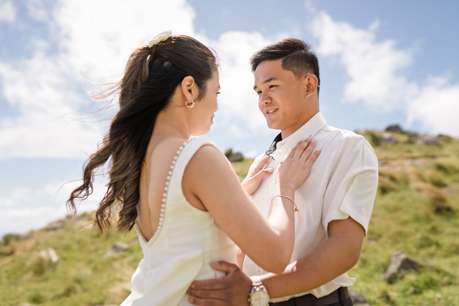 Couple in wedding attire sharing a romantic moment outdoors on a green hillside under a blue sky.