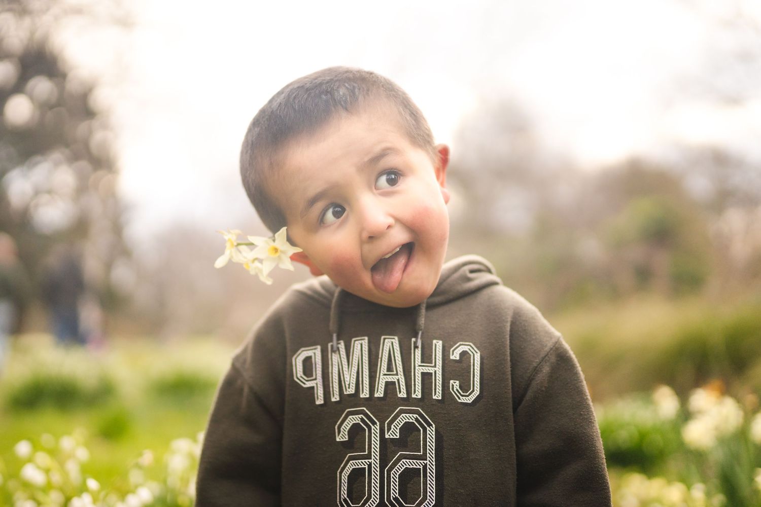 Cute toddler boy in a CHAMP hoodie sticking out his tongue outdoors among spring flowers.
