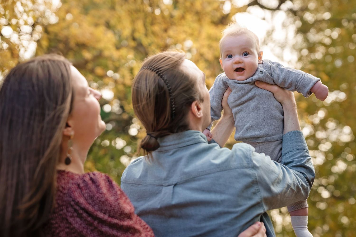 Two women holding a smiling baby outdoors in autumn park with golden foliage in the background.