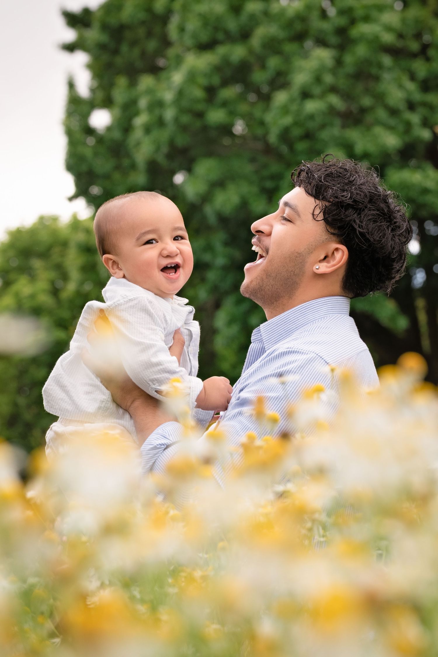 A joyful father laughs while holding his smiling baby outdoors among yellow wildflowers and green trees.