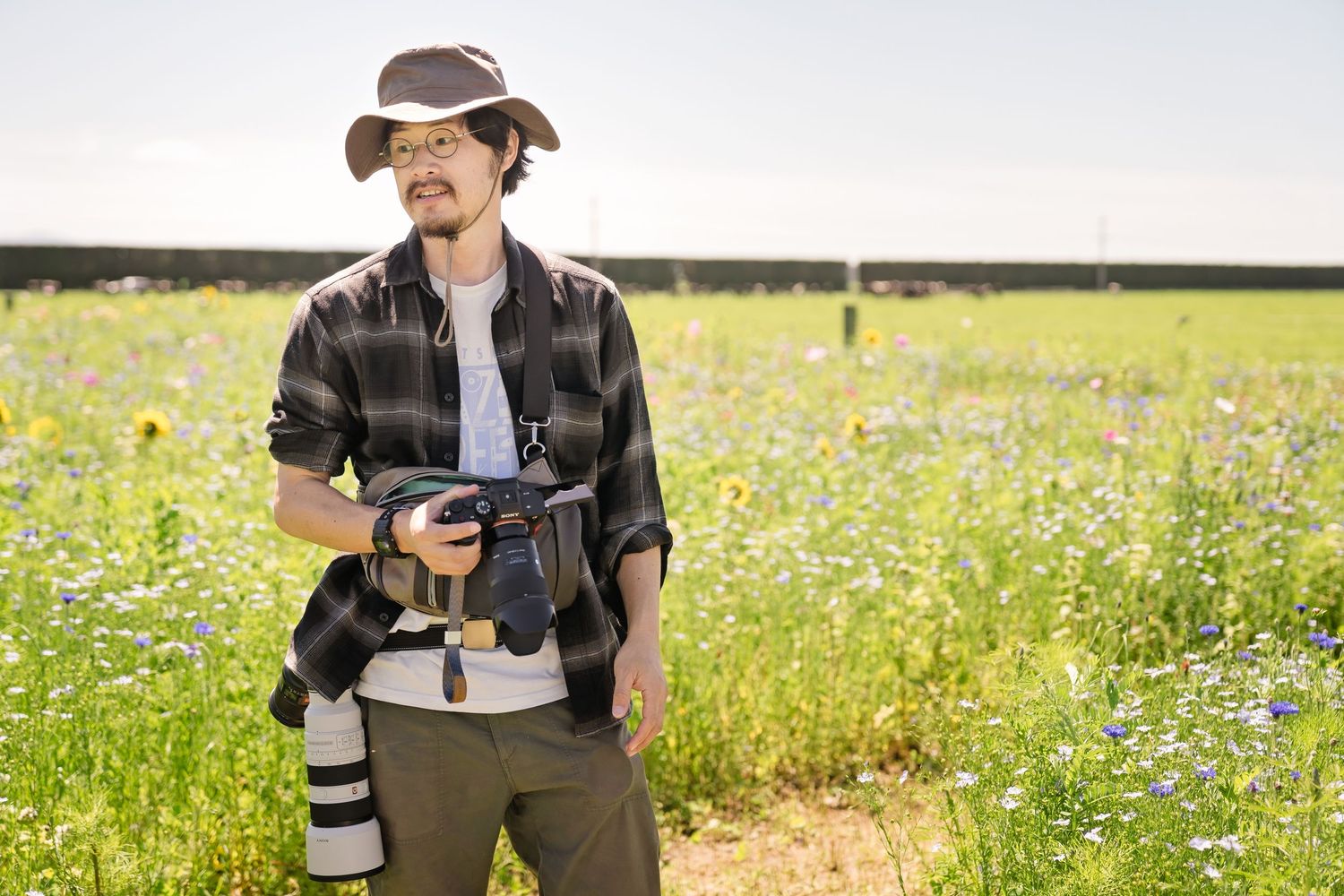Photographer in hat holding camera in colorful wildflower field on sunny day.