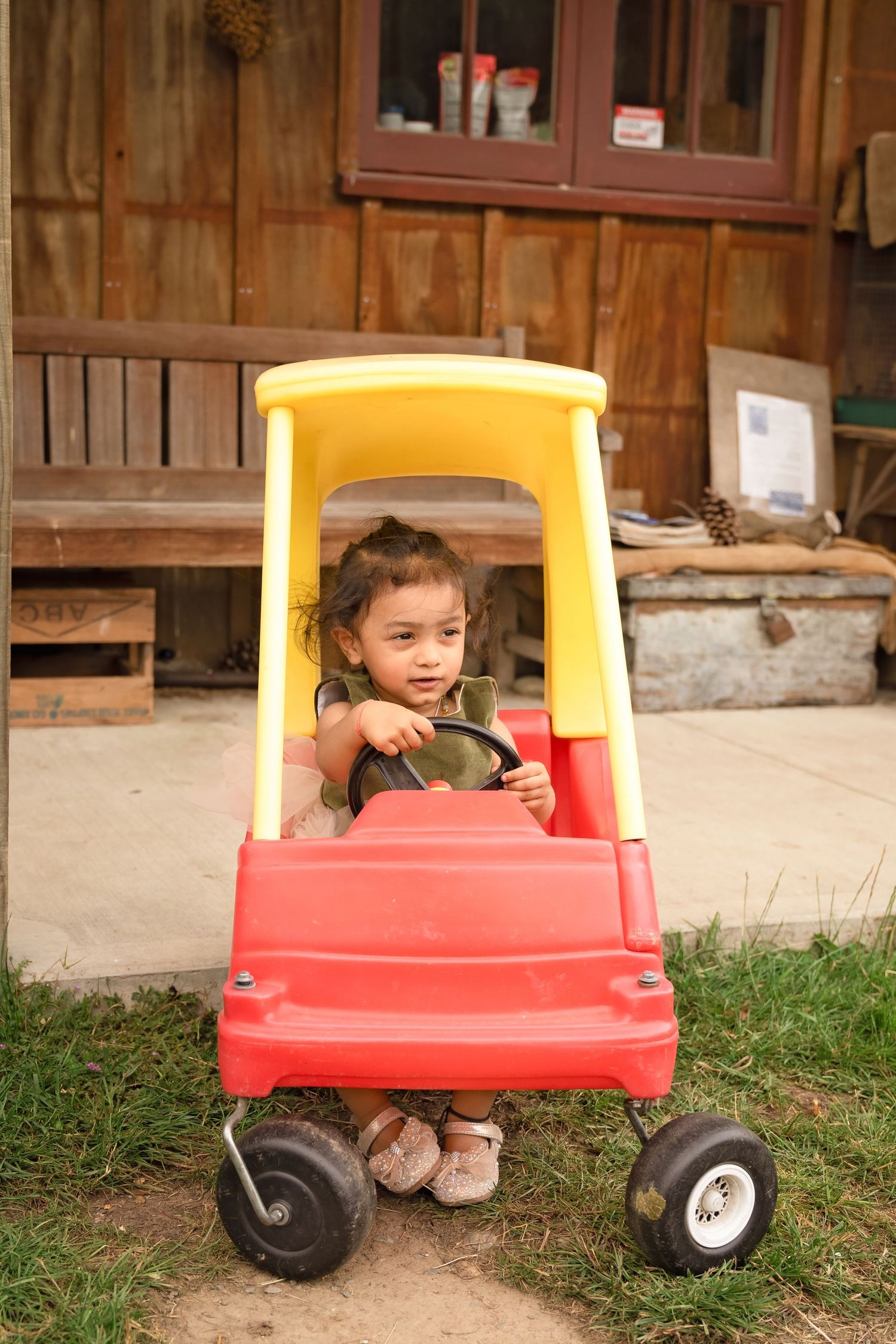 Child playing and hiding inside a red and yellow toy wagon on grass near a wooden barn.
