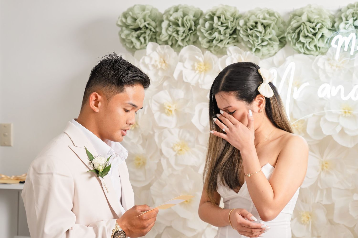 Emotional couple exchanging vows at wedding ceremony with white floral backdrop and neon sign.