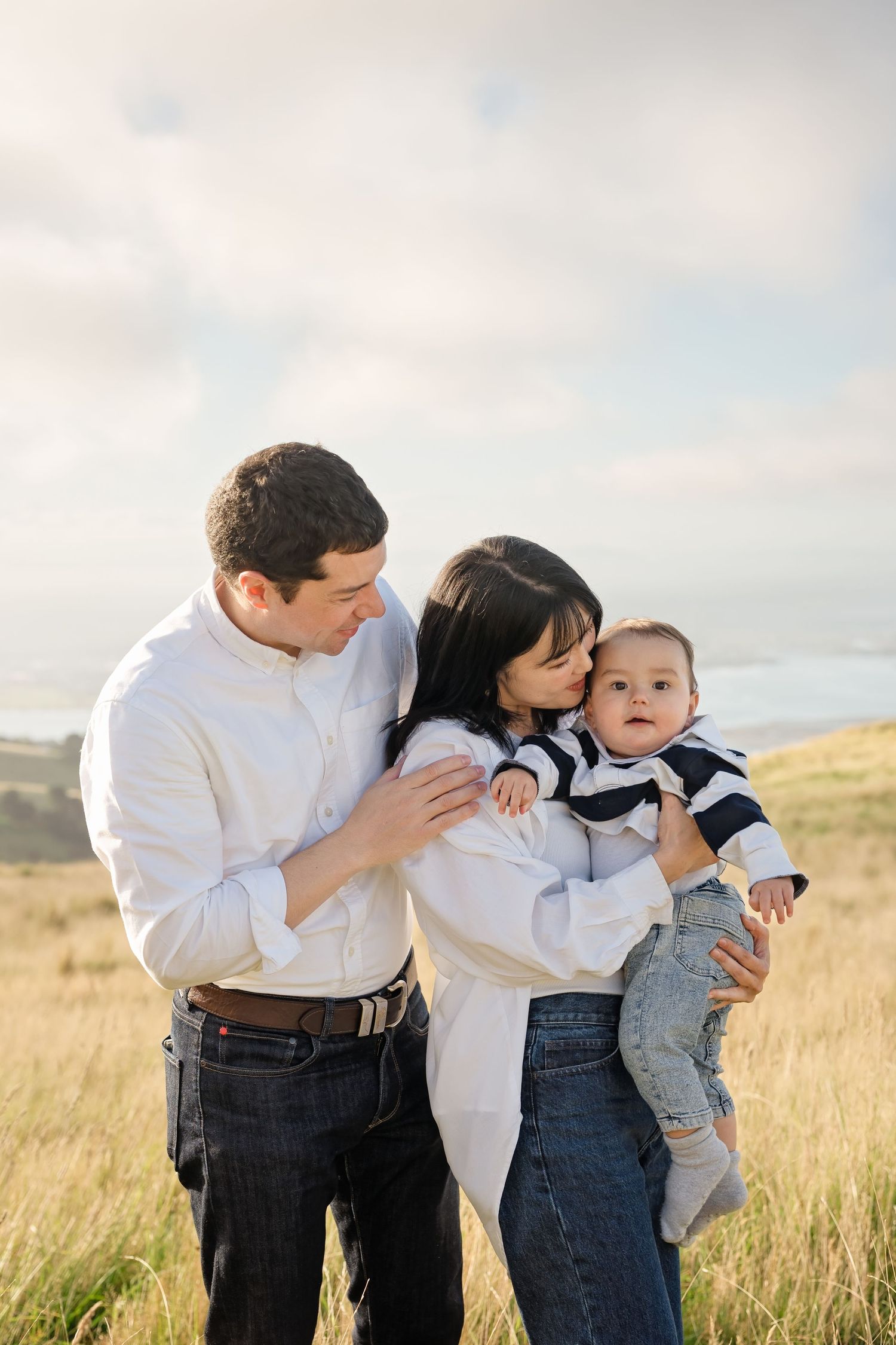 A young family poses outdoors in a grassy field, mother kissing baby while father looks on lovingly.