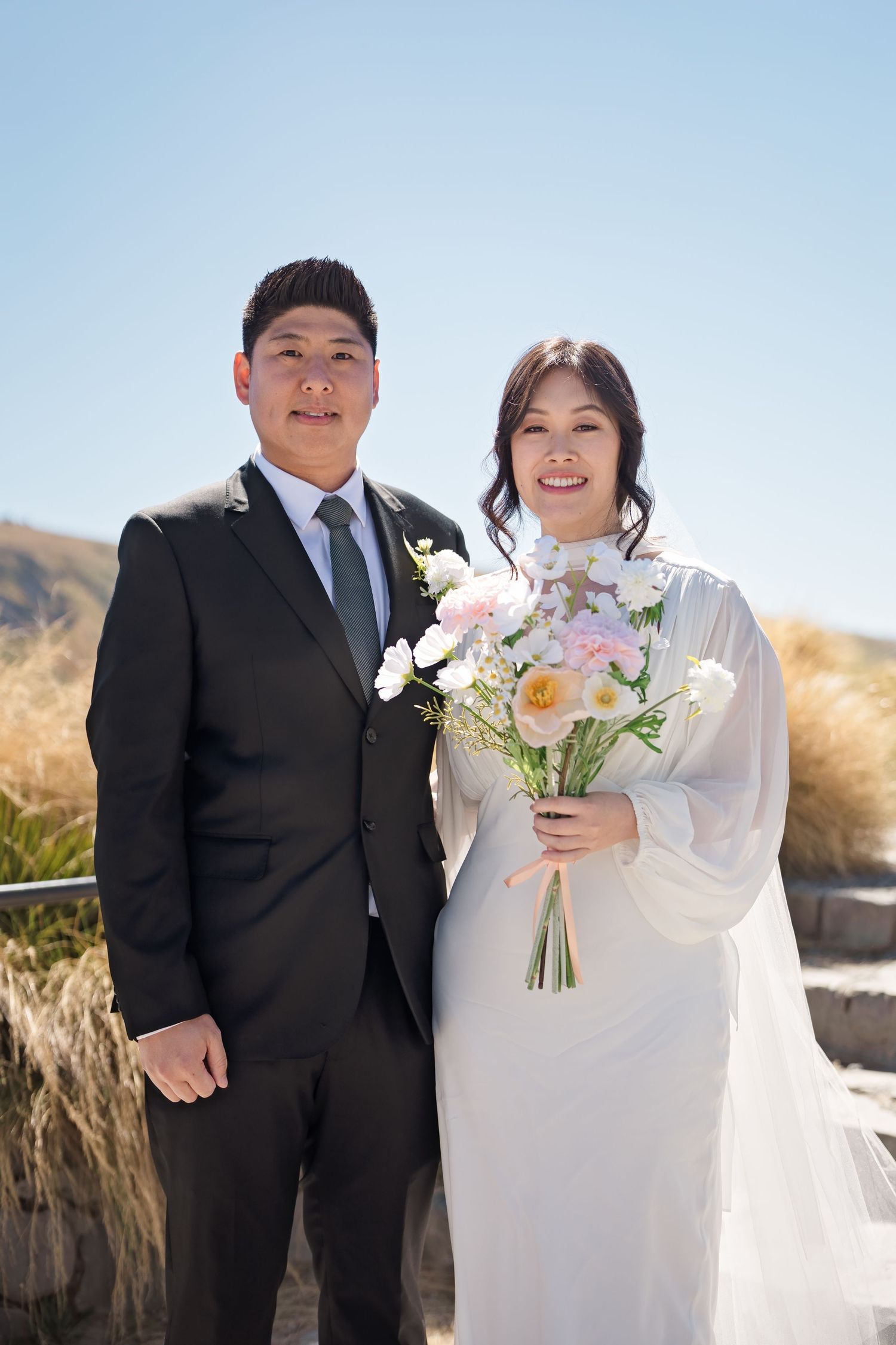 Smiling newlywed couple posing outdoors in wedding attire with bride holding floral bouquet.