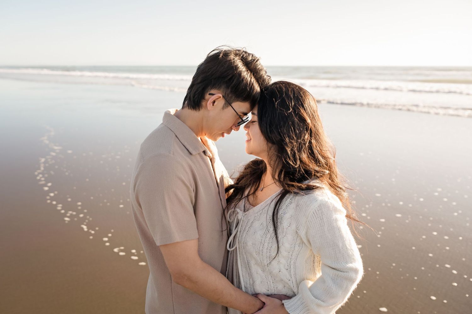 Romantic couple embracing on a sandy beach at sunset, touching foreheads in an intimate moment by the ocean shore.