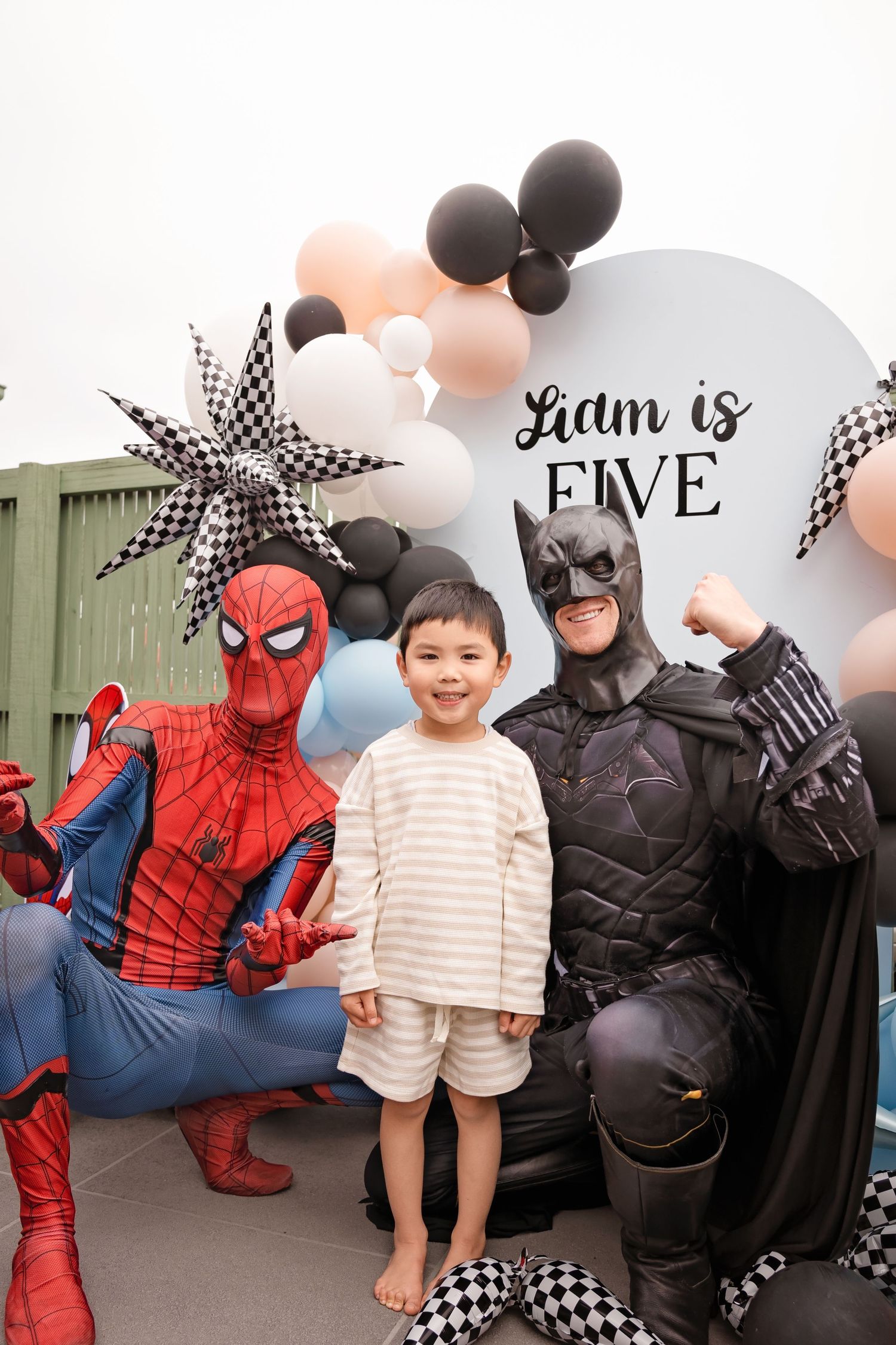 A young boy poses with Spider-Man and Batman costumed characters at Liam's fifth birthday party with balloon decorations.