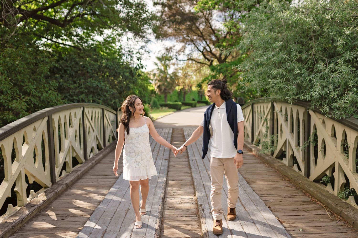 Couple holding hands walking on a wooden bridge surrounded by lush green trees on a sunny day.