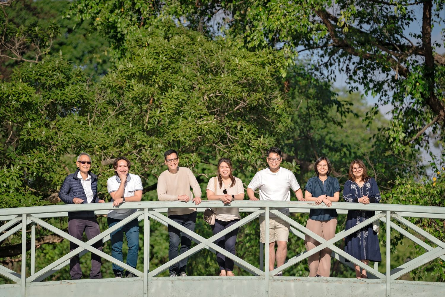A group of people stand together on a wooden bridge walkway surrounded by lush green foliage on a sunny day.