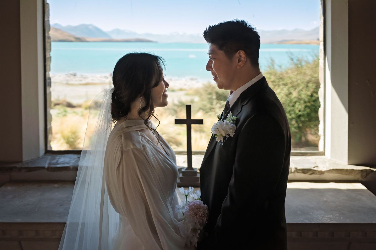 Bride and groom share an intimate moment before a wooden cross with turquoise lake views in a chapel.