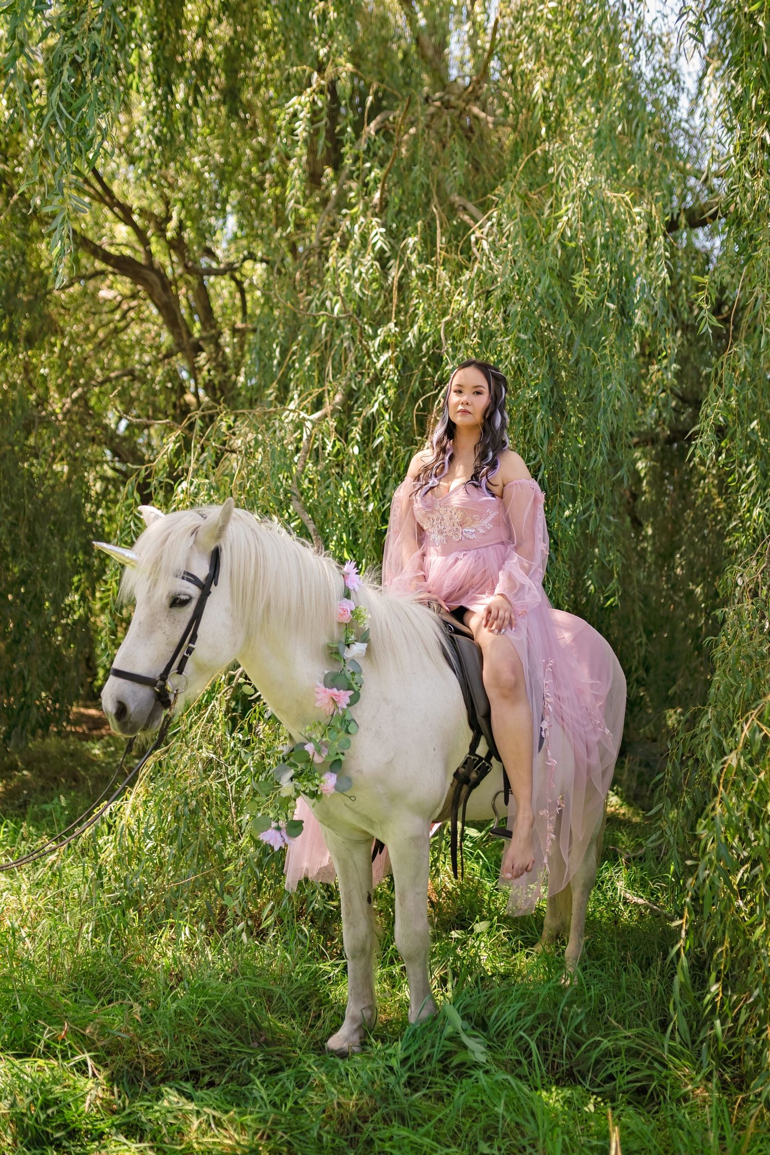 A woman in a pink gown rides a white horse decorated with flowers beneath weeping willow trees.