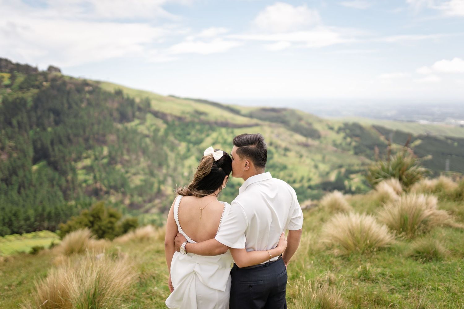 Bride and groom embracing on a lush green hilltop with panoramic valley views on their wedding day.