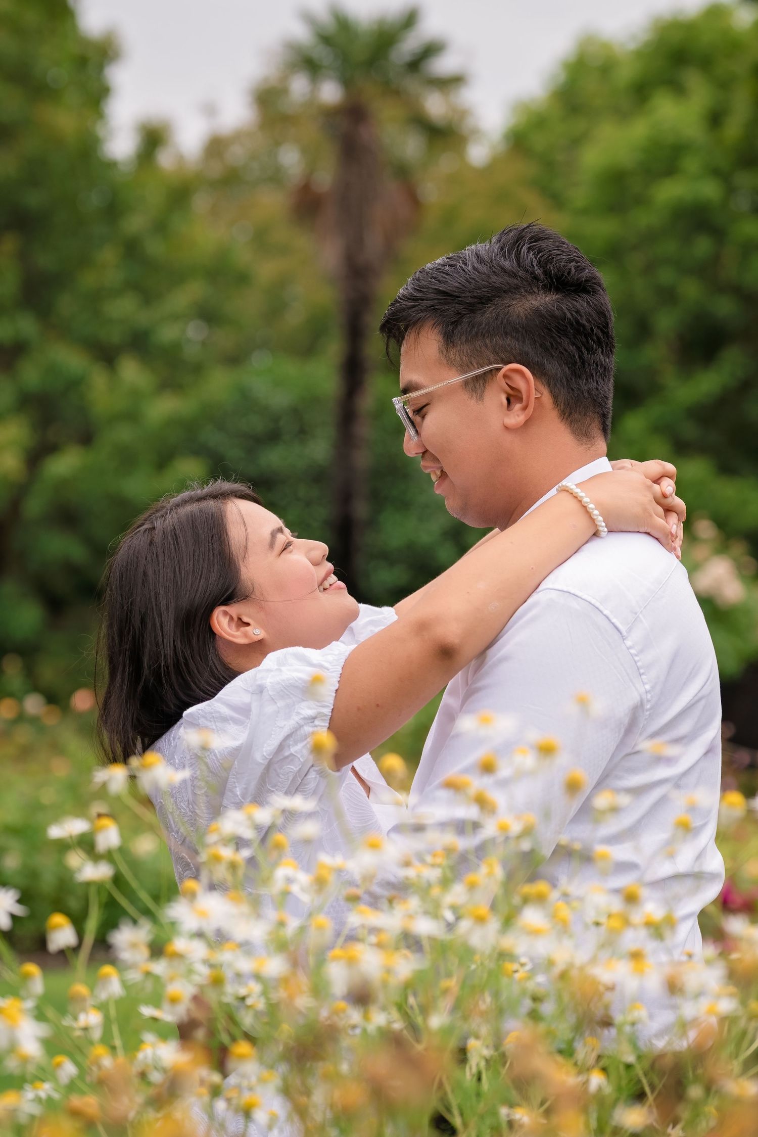 Couple embracing in a flower field, smiling at each other, surrounded by daisies and lush greenery.