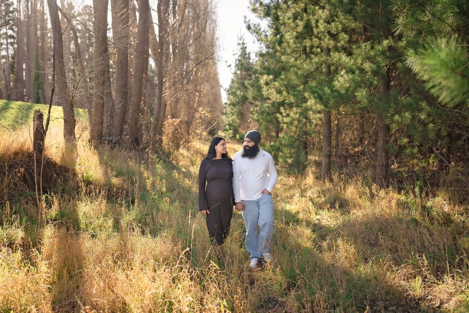 Couple walking hand in hand through a sunlit forest trail surrounded by tall pine trees and golden autumn grass.