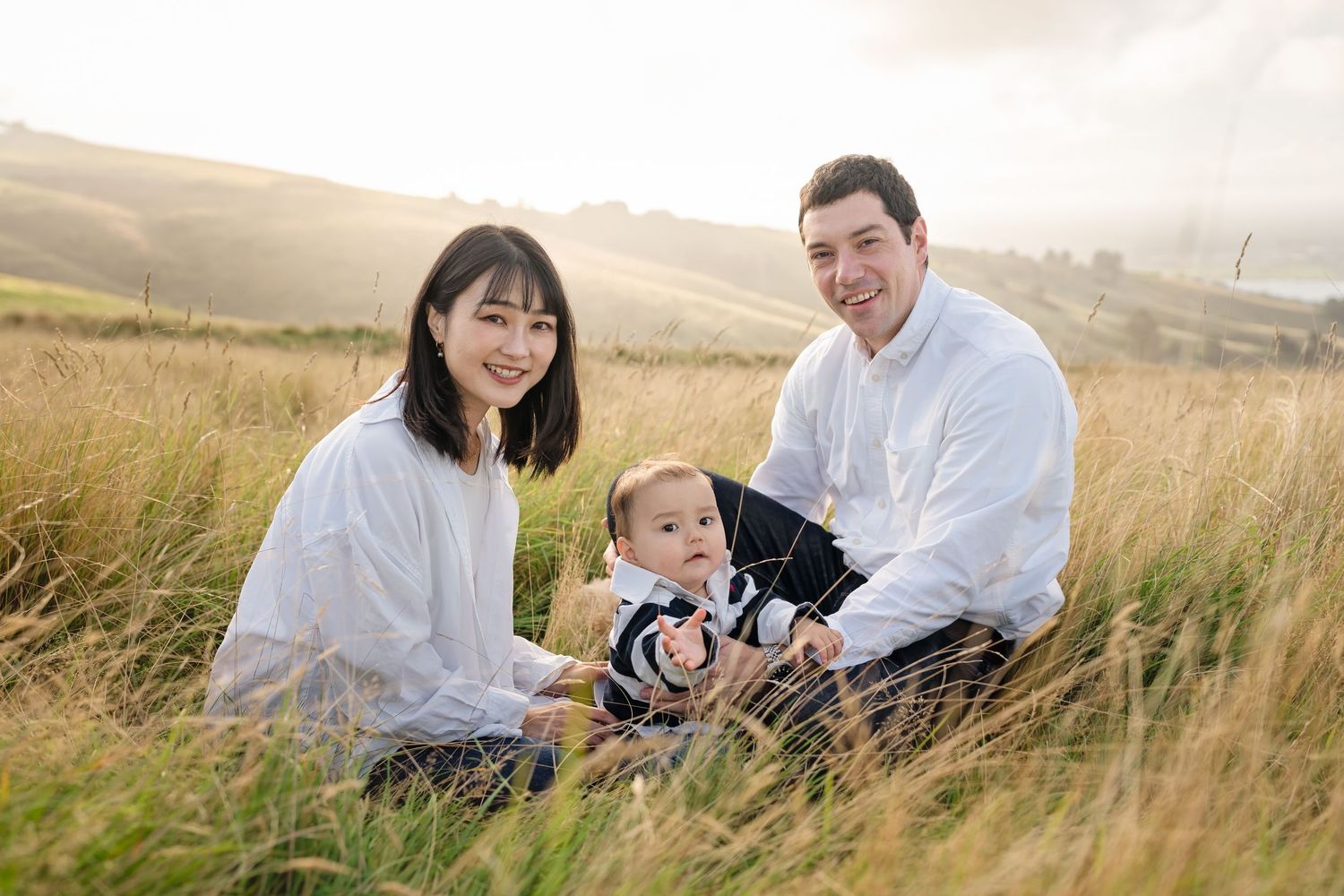 A smiling family of three sits in tall golden grass with rolling hills, parents posing with their baby boy outdoors.